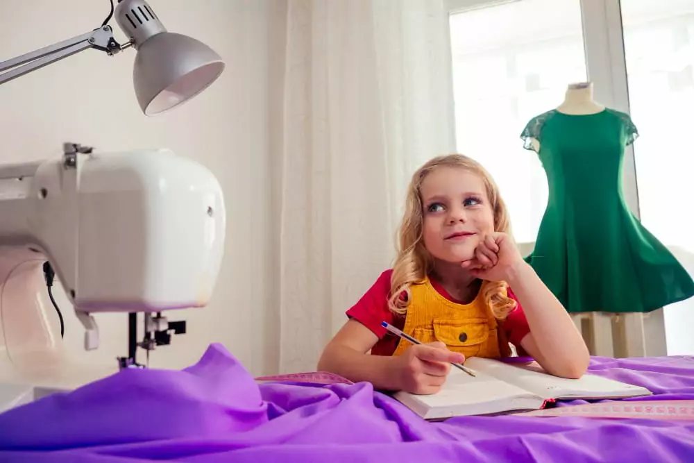 Lächelndes kleines blondes Mädchen am Tisch mit Nähmaschine und Schneiderpuppe