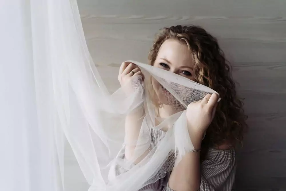 Ein lachendes junges Mädchen mit Locken, das am Fenster sitzt und einen weißen Vorhang in der Hand hält