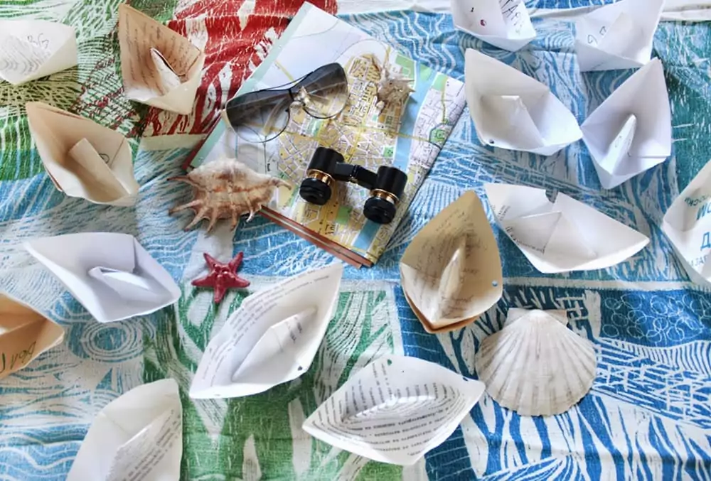 Papierboote, Muscheln, Fernglas und Karte auf buntem Stoff mit Sommermotiv.