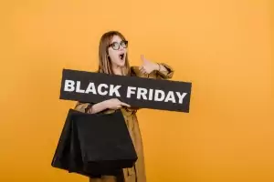 Lächelnde Frau mit Brille, Black Friday-Schild und Papiertüten vor gelbem Hintergrund